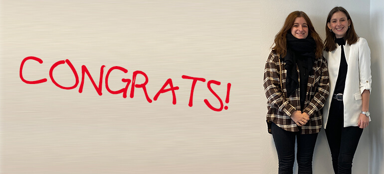 Zwei junge Frauen stehen vor einer Wand mit rotem Schriftzug CONGRATS und lächeln in die Kamera.
