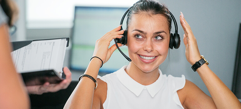 Frau setzt sich ein Headset auf und lächelt, Computerbildschirm im Hintergrund.
