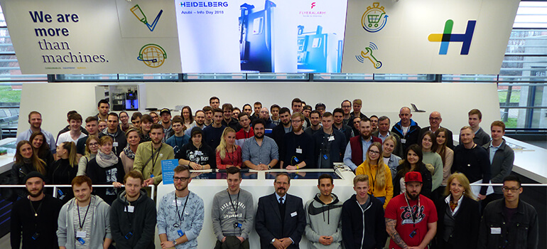 Gruppe von jungen Erwachsenen steht für ein Gruppenfoto vor Logos bei Heidelberg Druckmaschinen.