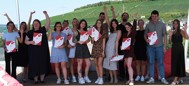 Gruppe junger Menschen mit Urkunden feiert draußen vor grüner Landschaft, einige halten die Arme hoch.