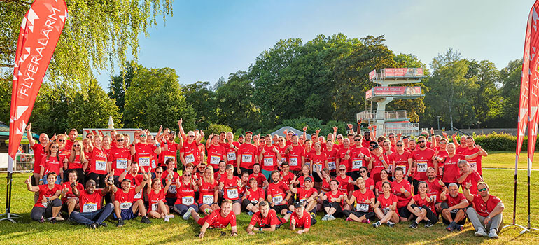 Große Gruppe in roten FLYERALARM-Shirts posiert im Grünen beim Firmenlauf.