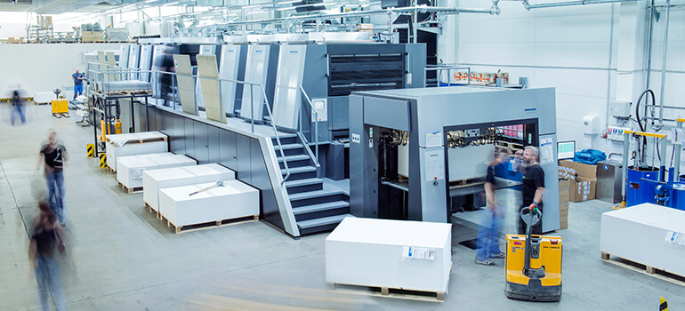 Große moderne Druckmaschine in einer Produktionshalle, Personen bewegen sich im Hintergrund.