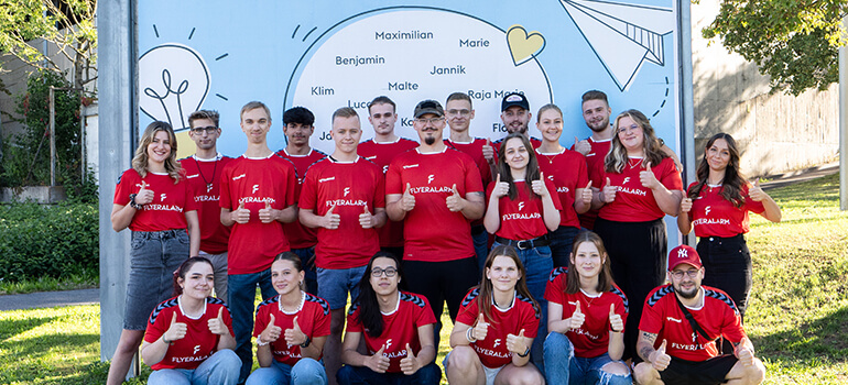 Gruppe junger Menschen in roten FLYERALARM-T-Shirts steht vor einem Plakat mit Namen, Herz und Papierflieger, viele zeigen den Daumen nach oben.