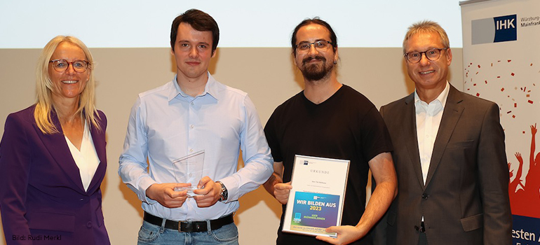Zwei Männer halten Urkunden und stehen mit einer Frau und einem Mann vor einem IHK-Aufsteller, alle lächeln in die Kamera.