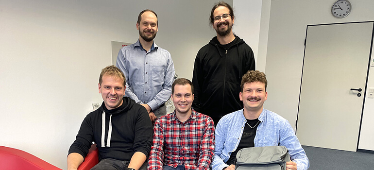 Fünf Männer sitzen und stehen lächelnd in einem Büroraum vor einer weißen Wand mit Tür und Uhr.