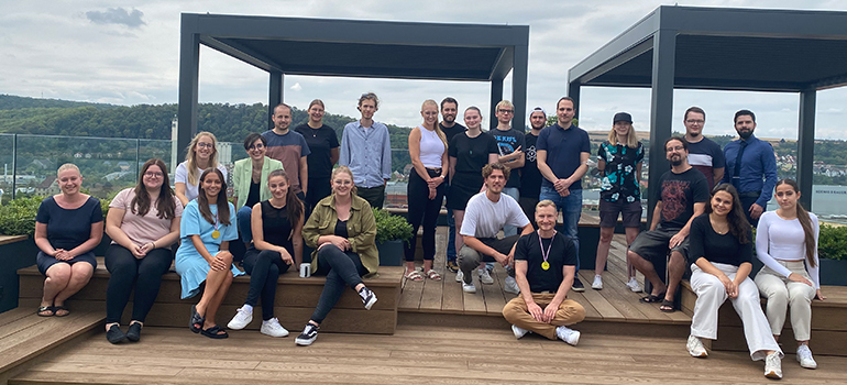 Gruppe junger Menschen mit Urkunden feiert draußen vor grüner Landschaft, einige halten die Arme hoch.