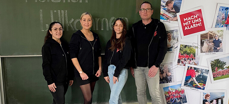 Vier Personen stehen in einem Klassenraum vor einer Tafel und einem Plakat mit FLYERALARM-Logo.