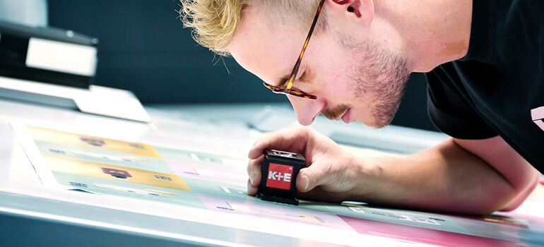 Mann prüft einen farbigen Druckbogen mit einer Lupe an einem Arbeitsplatz.